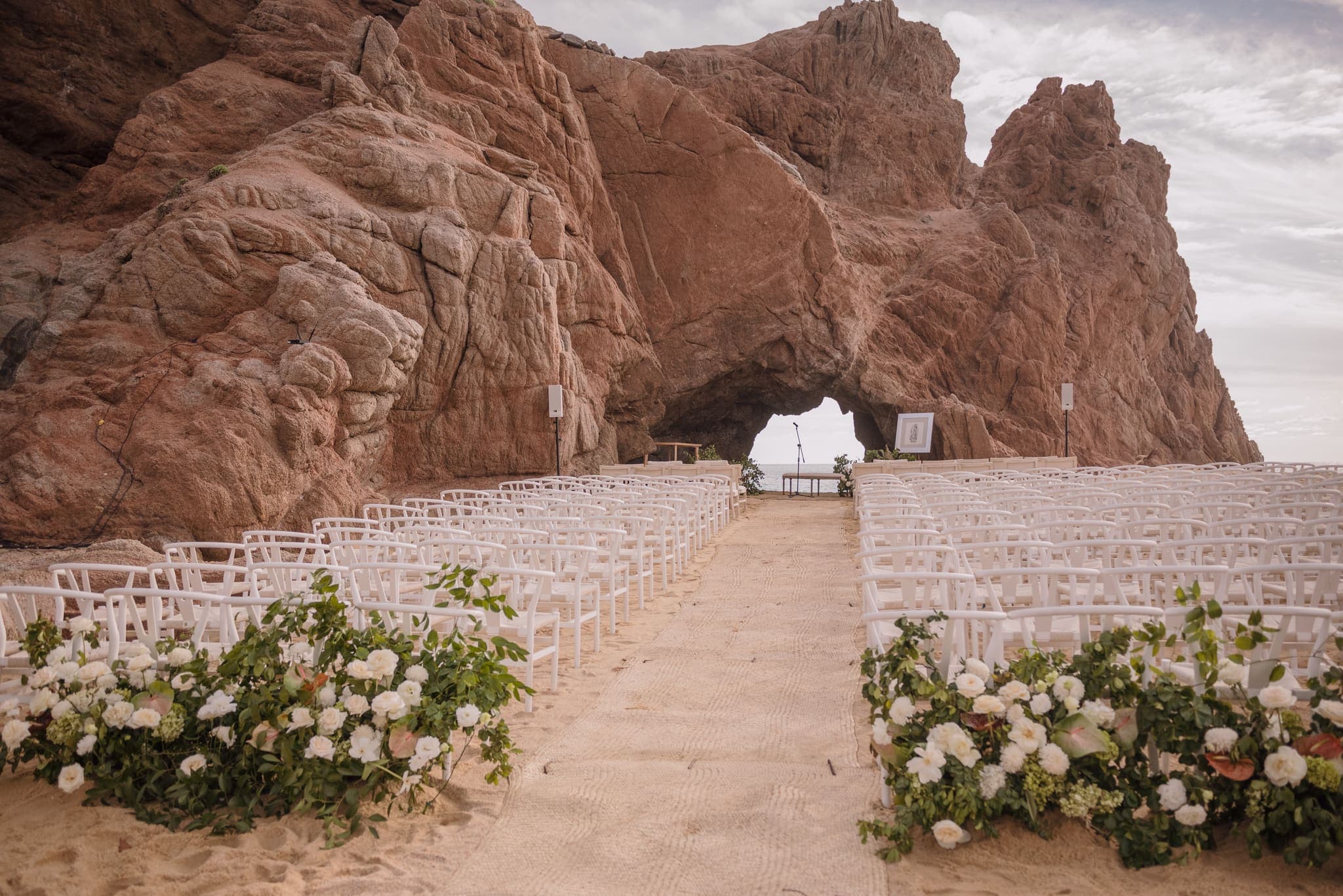 Ceremonia de boda frente a un arco de roca en Los Cabos, sillas blancas y flores