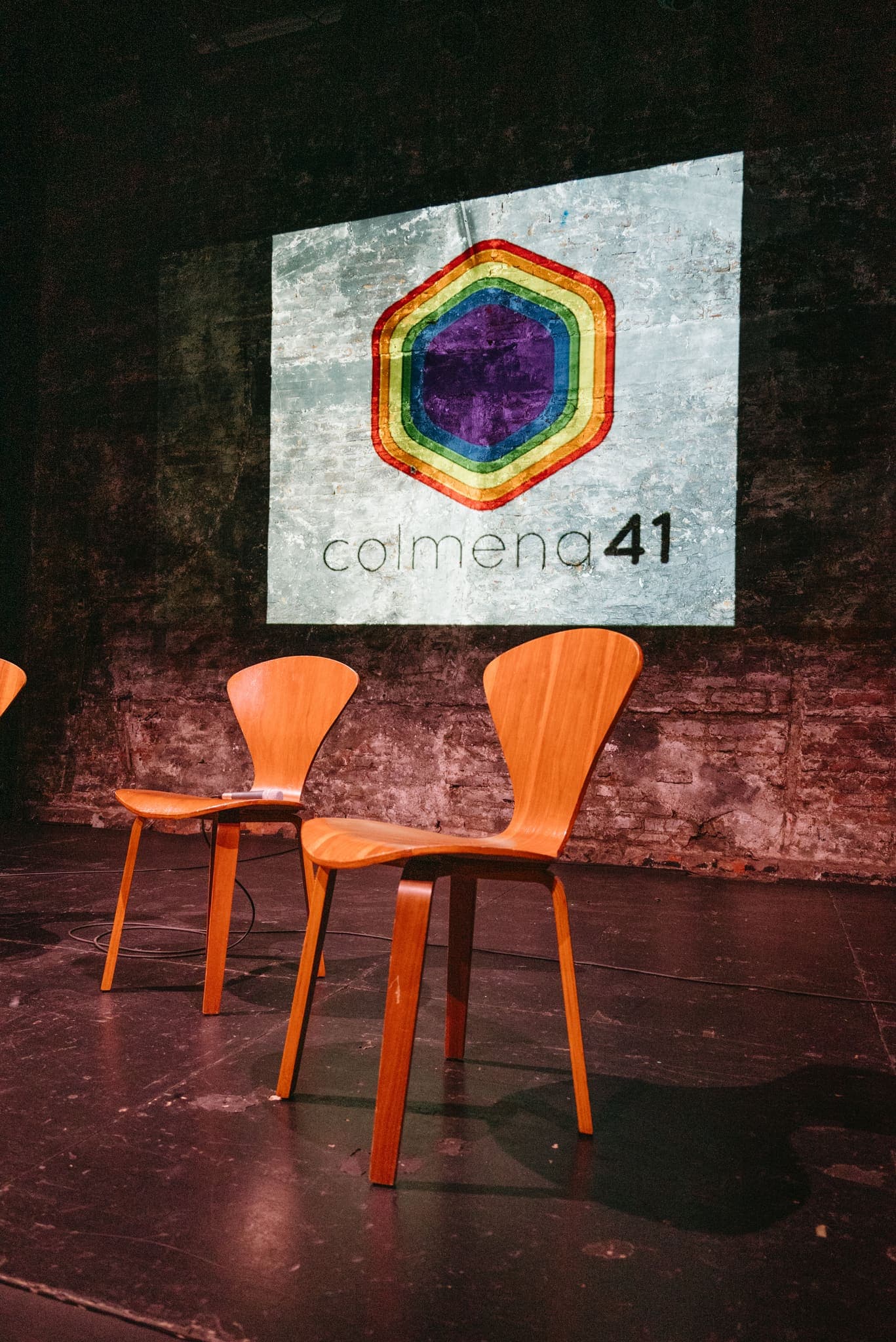 Escenario listo con sillas de madera bajo el logo de colmena41