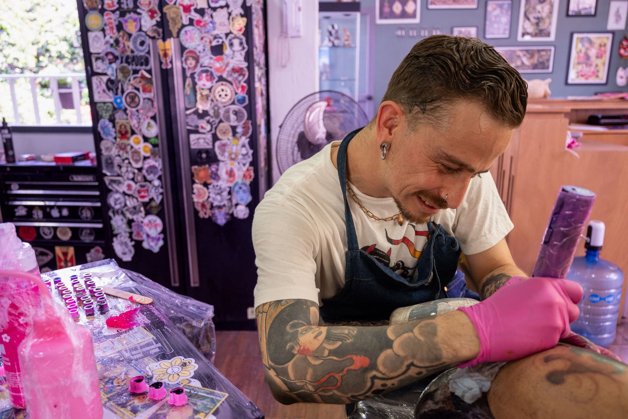 Tatuador sonriendo mientras trabaja, muro de calcomanías al fondo