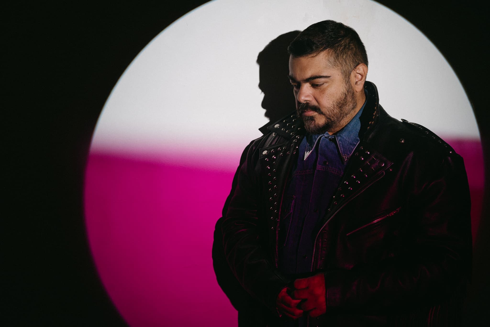 Retrato editorial con chaqueta de cuero bajo un círculo de luz rosa y blanca, cabeza inclinada